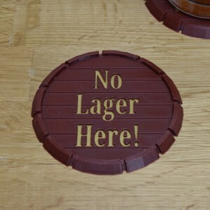 May include: A circular, brown coaster with the words "No Lager Here!" in gold lettering. The coaster is designed to resemble a barrel end, with horizontal lines and a rounded edge. The coaster is on a wooden surface.