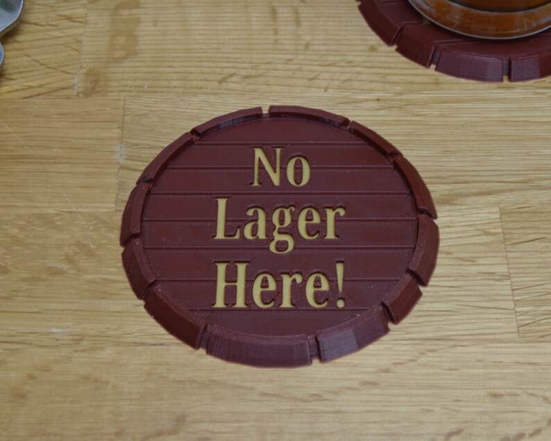 May include: A circular, brown coaster with the words "No Lager Here!" in gold lettering. The coaster is designed to resemble a barrel end, with horizontal lines and a rounded edge. The coaster is on a wooden surface.