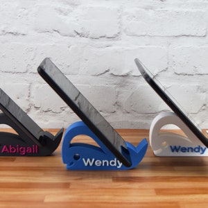 May include: Three phone stands shaped like whales, each with a different color. The stands are holding phones in a vertical position. The stands are labeled "Abigail", "Wendy", and "Wendy".