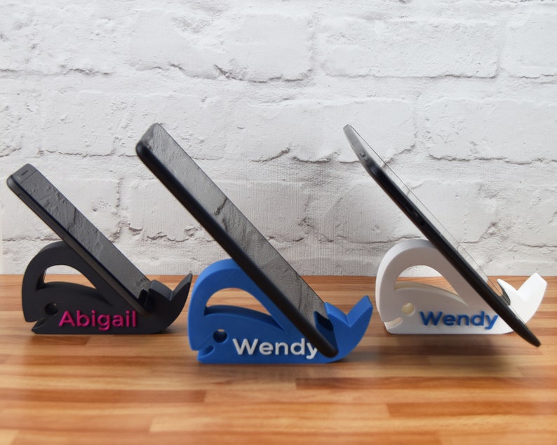 May include: Three phone stands shaped like whales, each with a different color. The stands are holding phones in a vertical position. The stands are labeled "Abigail", "Wendy", and "Wendy".