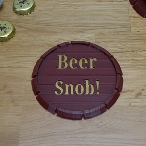 May include: A round, brown, wooden coaster with the words "Beer Snob!" in gold lettering.