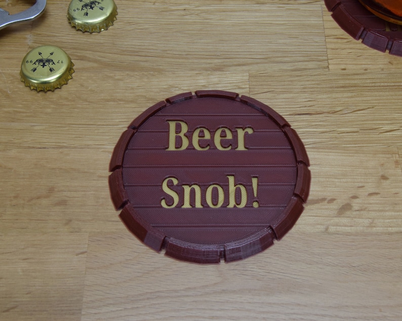 May include: A round, brown, wooden coaster with the words "Beer Snob!" in gold lettering.