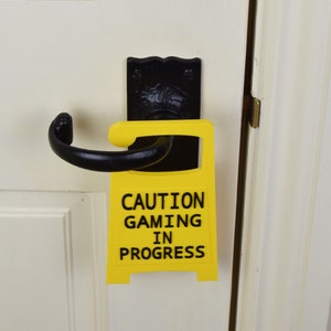 May include: A yellow sign with black text that reads "Caution Gaming In Progress" is attached to a black door handle. The sign is a fun way to let people know that you are busy gaming.