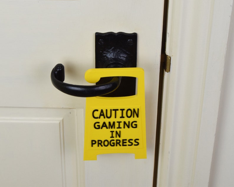 May include: A yellow sign with black text that reads "Caution Gaming In Progress" is attached to a black door handle. The sign is a fun way to let people know that you are busy gaming.
