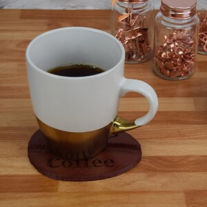 May include: A white coffee mug with a gold rim and a brown leather coaster with the word "Coffee" engraved on it. The mug is filled with coffee.