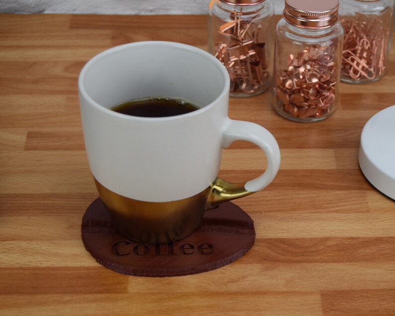 May include: A white coffee mug with a gold rim and a brown leather coaster with the word "Coffee" engraved on it. The mug is filled with coffee.
