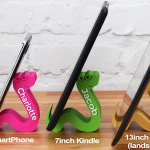 May include: Three colorful 3D printed snake phone and tablet stands. The pink stand is holding a 6 inch smartphone and says "Charlotte". The green stand is holding a 7 inch Kindle and says "Jacob". The gold stand is holding a 13 inch tablet in landscape mode and says "Oliver".