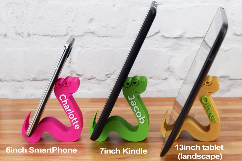 May include: Three colorful 3D printed snake phone and tablet stands. The pink stand is holding a 6 inch smartphone and says "Charlotte". The green stand is holding a 7 inch Kindle and says "Jacob". The gold stand is holding a 13 inch tablet in landscape mode and says "Oliver".