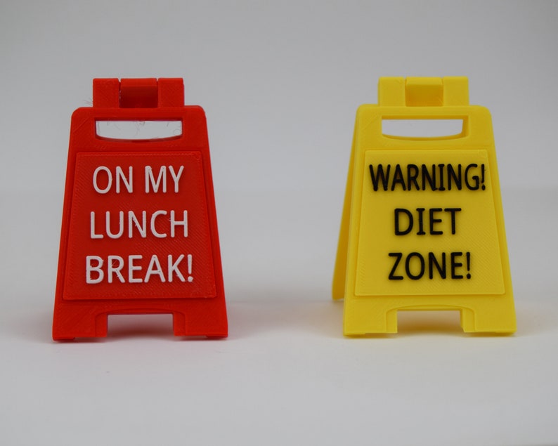 May include: Two small, plastic sign props. One is red and reads "ON MY LUNCH BREAK!" in white. The other is yellow and reads "WARNING! DIET ZONE!" in black. Both signs are shaped like caution signs.