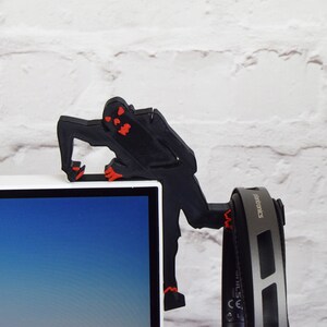 May include: A black 3D printed figure of a monster with red eyes climbing on a white computer monitor. The figure is holding a pair of black headphones with the word "electronics" printed on the side.