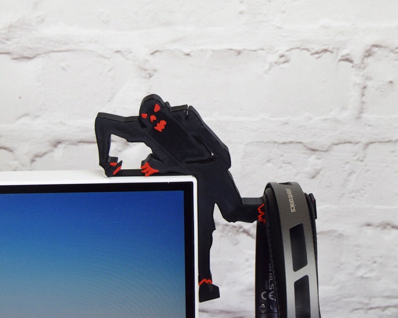 May include: A black 3D printed figure of a monster with red eyes climbing on a white computer monitor. The figure is holding a pair of black headphones with the word "electronics" printed on the side.