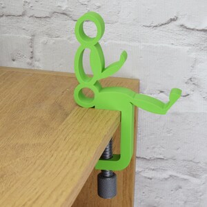 May include: A green plastic figure of a person sitting on a wooden surface. The figure is attached to a clamp that is used to hold the figure in place.