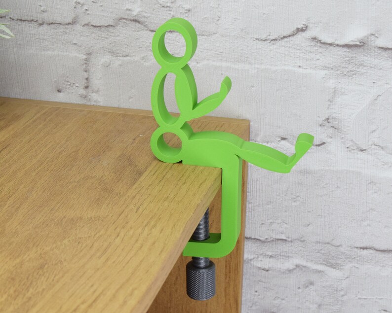 May include: A green plastic figure of a person sitting on a wooden surface. The figure is attached to a clamp that is used to hold the figure in place.