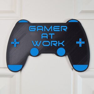 以下が含まれることがあります： 黒と青のゲームコントローラーのサインで、「Gamer at Work」というテキストが書かれています。
