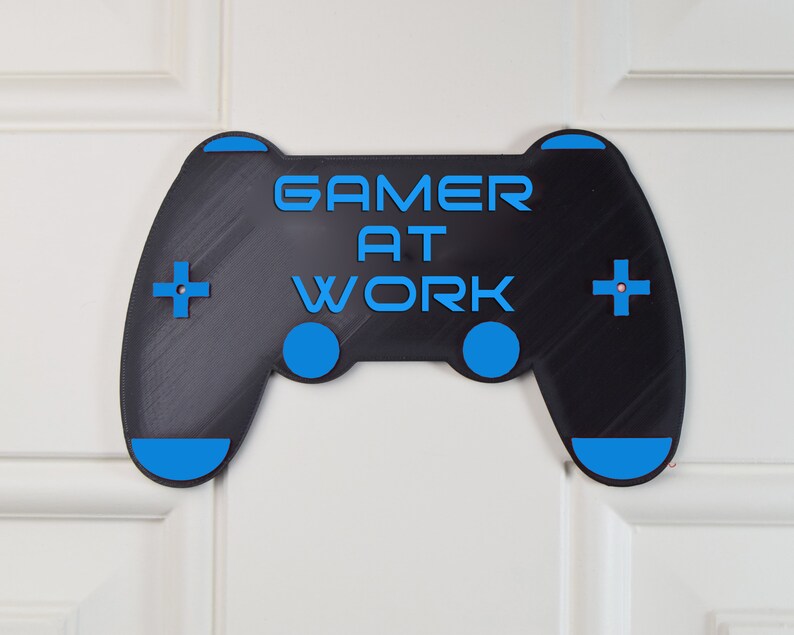 以下が含まれることがあります： 黒と青のゲームコントローラーのサインで、「Gamer at Work」というテキストが書かれています。