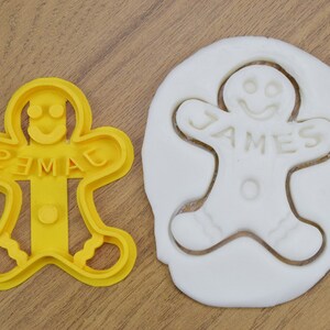 Puede incluir: Cortador de galletas de jengibre amarillo en forma de hombre con el nombre "JAMES" escrito en &eacute;l. Una masa de galletas de jengibre blanca se presiona en el cortador.