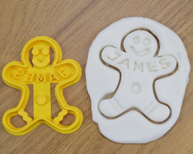 Puede incluir: Cortador de galletas de jengibre amarillo en forma de hombre con el nombre "JAMES" escrito en &eacute;l. Una masa de galletas de jengibre blanca se presiona en el cortador.
