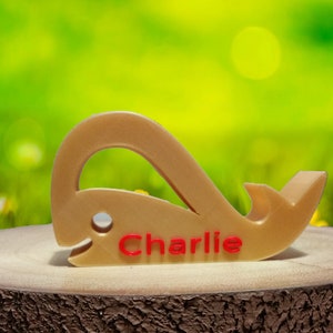 May include: A wooden whale-shaped doorstop with the name "Charlie" engraved in red lettering. The doorstop is sitting on a wooden surface.