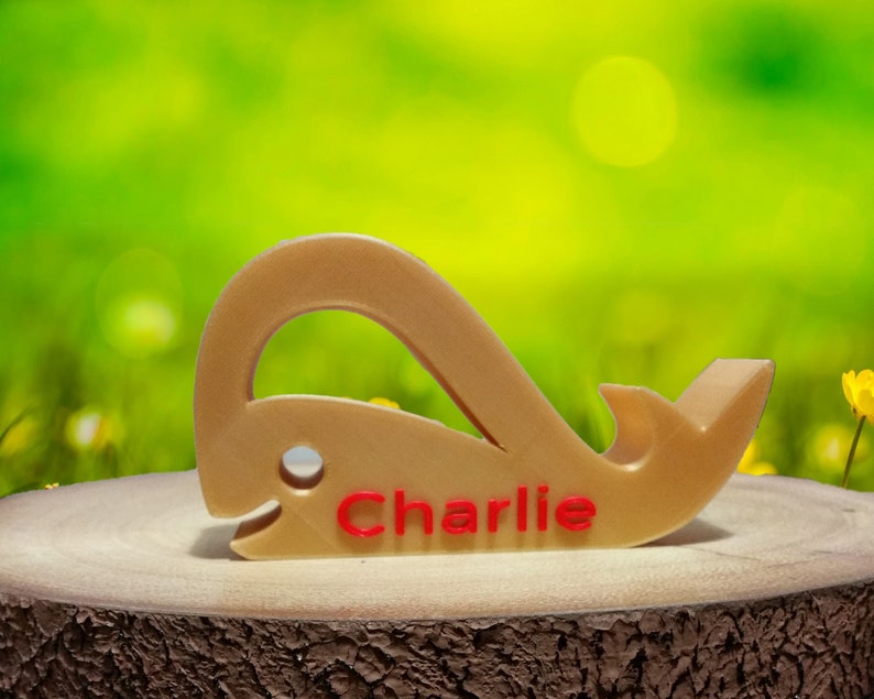 May include: A wooden whale-shaped doorstop with the name "Charlie" engraved in red lettering. The doorstop is sitting on a wooden surface.