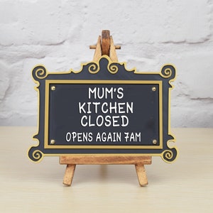 May include: Black sign with gold trim and ornate edges on a wooden easel. The sign reads "Mum's Kitchen Closed Opens Again 7am".