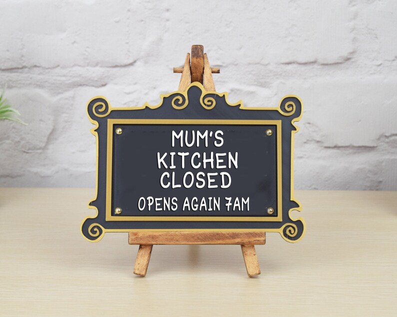 May include: Black sign with gold trim and ornate edges on a wooden easel. The sign reads "Mum's Kitchen Closed Opens Again 7am".