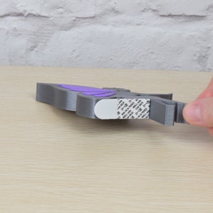 K&ouml;nnte beinhalten: Eine graue, 3D-gedruckte Schmetterlings-Wanddekoration mit violetten Fl&uuml;geln. Der Schmetterling hat Klebeband auf der R&uuml;ckseite zum einfachen Aufh&auml;ngen.