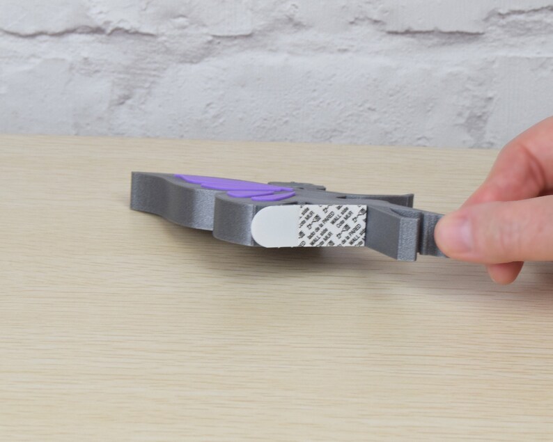K&ouml;nnte beinhalten: Eine graue, 3D-gedruckte Schmetterlings-Wanddekoration mit violetten Fl&uuml;geln. Der Schmetterling hat Klebeband auf der R&uuml;ckseite zum einfachen Aufh&auml;ngen.