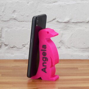 K&ouml;nnte beinhalten: Ein rosa Pinguin-Handyst&auml;nder mit dem Namen "Angela" auf der Seite. Der St&auml;nder h&auml;lt ein schwarzes Smartphone.