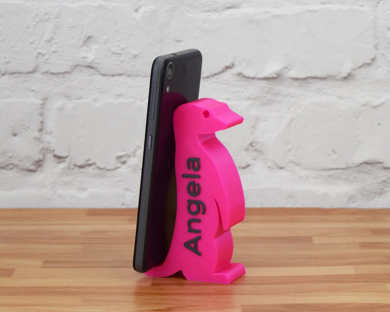 K&ouml;nnte beinhalten: Ein rosa Pinguin-Handyst&auml;nder mit dem Namen "Angela" auf der Seite. Der St&auml;nder h&auml;lt ein schwarzes Smartphone.