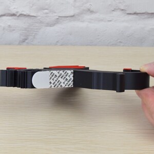 May include: A black, 3D-printed object with red accents and a white adhesive pad. The object is held by a hand and appears to be a wall mount or organizer. The object is on a light wood surface.