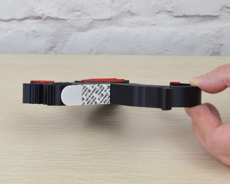 May include: A black, 3D-printed object with red accents and a white adhesive pad. The object is held by a hand and appears to be a wall mount or organizer. The object is on a light wood surface.