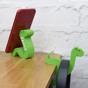 May include: Two green snake-shaped phone stands. One stand is holding a red smartphone with the name "Jacob" printed on it. The other stand is holding a black headphone.