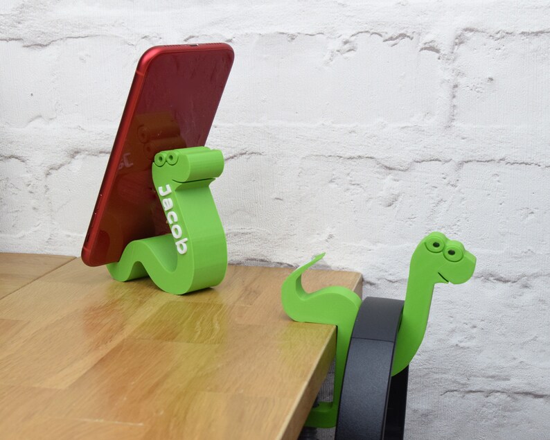May include: Two green snake-shaped phone stands. One stand is holding a red smartphone with the name "Jacob" printed on it. The other stand is holding a black headphone.