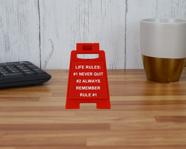 May include: A red sign with the text "LIFE RULES: #1 NEVER QUIT #2 ALWAYS REMEMBER RULE #1" stands on a wooden desk. A black keyboard and a white and gold mug are in the background.