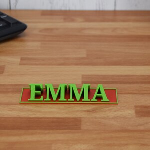 May include: A green 3D-printed nameplate with the name "EMMA" in capital letters. The letters are raised and sit on a red rectangular base. The nameplate is on a wooden desk.