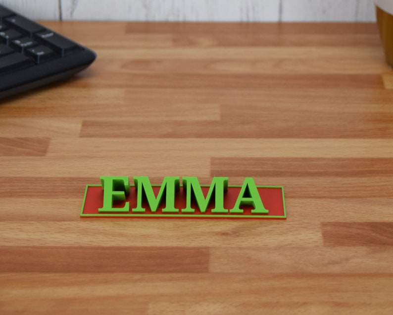 May include: A green 3D-printed nameplate with the name "EMMA" in capital letters. The letters are raised and sit on a red rectangular base. The nameplate is on a wooden desk.