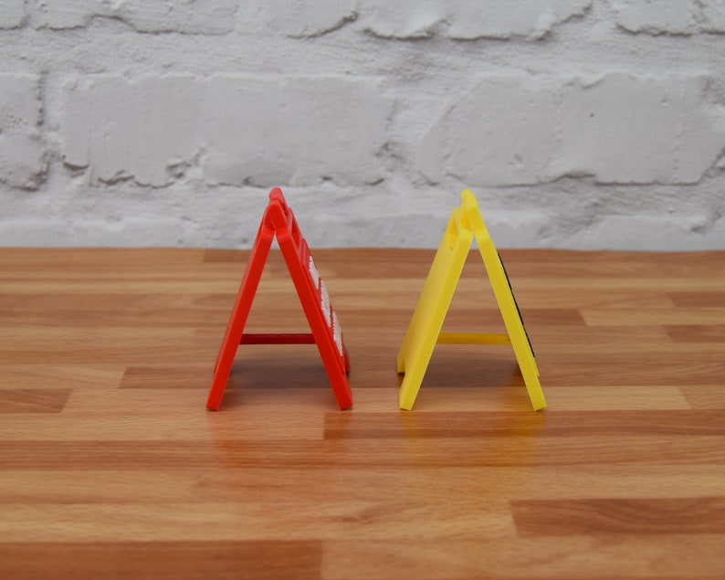May include: Two foldable road signs, one red and one yellow, are standing upright on a wooden surface. The signs are made of plastic and have a triangular shape.