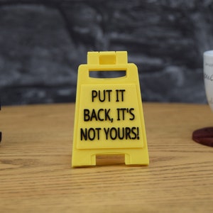 May include: A yellow plastic sign with the text "PUT IT BACK, IT'S NOT YOURS!" in black. The sign is in a triangular shape, designed to stand upright. A stapler and a coffee cup are in the background.
