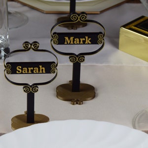 May include: Two black metal table card holders with gold accents. The card holders have a gold base and a black stand. The cards are black with gold lettering. The cards read "Mark" and "Sarah".