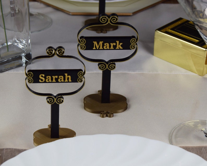 May include: Two black metal table card holders with gold accents. The card holders have a gold base and a black stand. The cards are black with gold lettering. The cards read "Mark" and "Sarah".