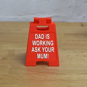 May include: A red plastic sign with the text "DAD IS WORKING ASK YOUR MUM!" on it.