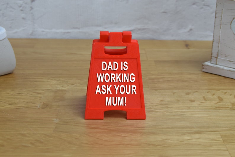 May include: A red plastic sign with the text "DAD IS WORKING ASK YOUR MUM!" on it.