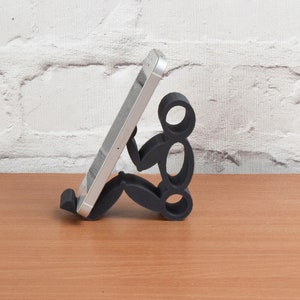 May include: A black phone stand shaped like a person sitting down, holding a silver smartphone.
