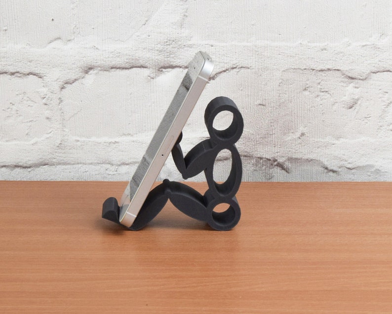May include: A black phone stand shaped like a person sitting down, holding a silver smartphone.