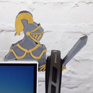 Op de afbeelding: Een grijs 3D-geprint ridderfiguur met een gouden helm en harnas, dat een zwaard vasthoudt, zit bovenop een computermonitor. De monitor is zwart en heeft een blauw scherm. De ridder houdt een paar zwarte koptelefoons vast met het woord "Plantronics" erop gedrukt.