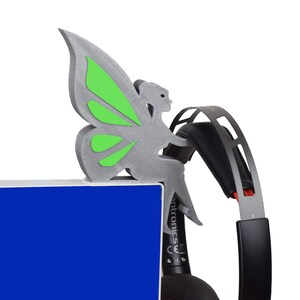 K&ouml;nnte beinhalten: Eine silberne, 3D-gedruckte Feenfigur mit gr&uuml;nen Fl&uuml;geln, die auf einem schwarzen Headset sitzt. Das Headset liegt auf einer blauen Oberfl&auml;che.
