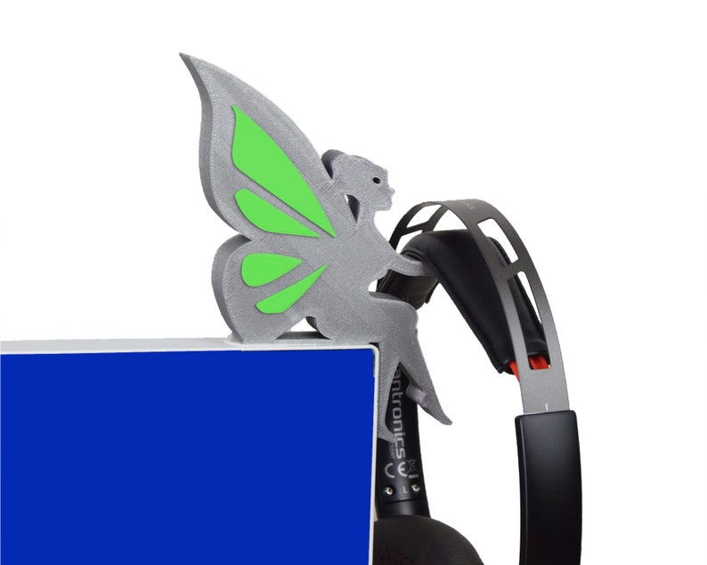 K&ouml;nnte beinhalten: Eine silberne, 3D-gedruckte Feenfigur mit gr&uuml;nen Fl&uuml;geln, die auf einem schwarzen Headset sitzt. Das Headset liegt auf einer blauen Oberfl&auml;che.