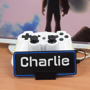 Pu&ograve; includere: Un controller per videogiochi bianco con pulsanti neri &egrave; posizionato su un supporto nero con un bordo al neon blu. Il supporto ha la scritta "Charlie" stampata in bianco.