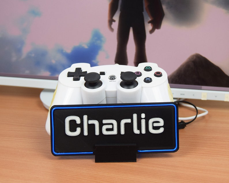 Pu&ograve; includere: Un controller per videogiochi bianco con pulsanti neri &egrave; posizionato su un supporto nero con un bordo al neon blu. Il supporto ha la scritta "Charlie" stampata in bianco.