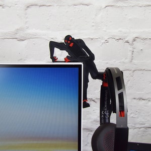 May include: A black 3D printed figure of a monster with red eyes is holding a pair of black headphones with red accents. The headphones have the text "Plantronics" on the side. The figure is perched on the top of a black computer monitor.